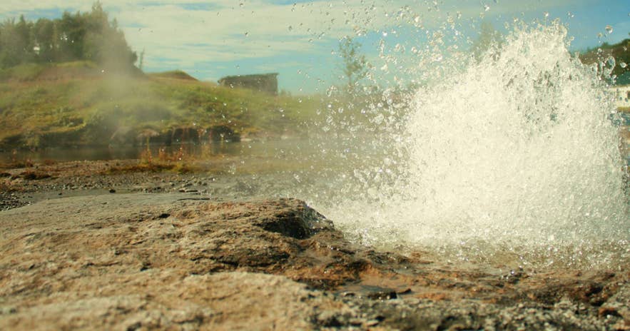 Litli Geysir spraying hot water across rocky ground, with the old stone building at the Secret Lagoon behind