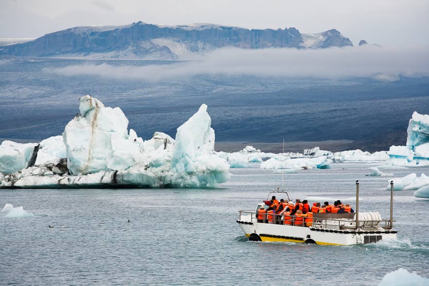 Toeristen met oranje reddingsvesten op een amfibieboot tussen drijvende ijsbergen op de Jokulsarlon-gletsjerlagune in Zuidoost-IJsland, met mistige bergen en gletsjerlandschap op de achtergrond.