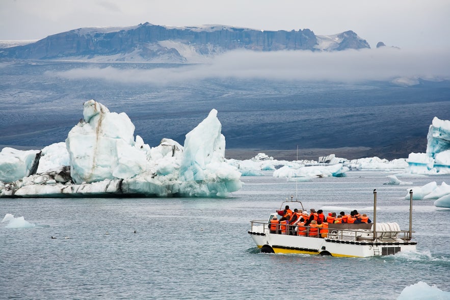 Turistas con chalecos salvavidas naranjas en un tour en barco anfibio entre icebergs flotantes en la laguna glaciar de J&ouml;kuls&aacute;rl&oacute;n en el sureste de Islandia, con monta&ntilde;as y glaciares al fondo.