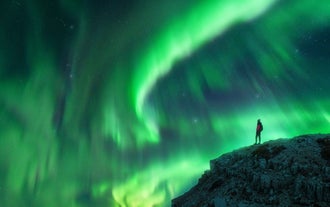 A person stands on a rocky hill in Iceland, admiring the vivid green northern lights dancing across the night sky.