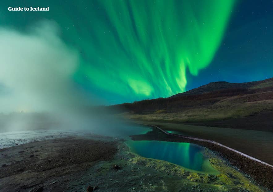 Nordlys over geotermisk varm kilde på Island med damp, fargerik aurora og vulkansk landskap om natten