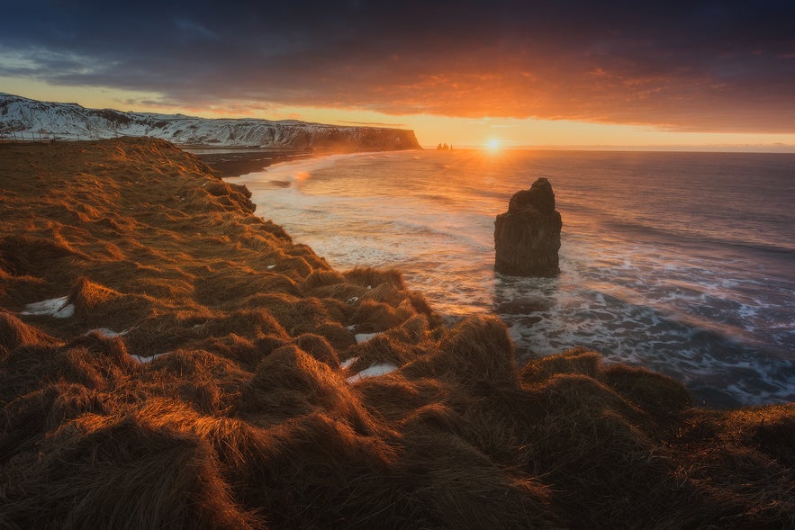 Dramatic sunset over the South Coast of Iceland with waves crashing against sea stacks and golden light on the coastal cliffs near Vik.
