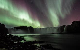 Nordlys i grønne og lilla nuancer lyser op over Godafoss-vandfaldet, der bruser i nattens mørke.