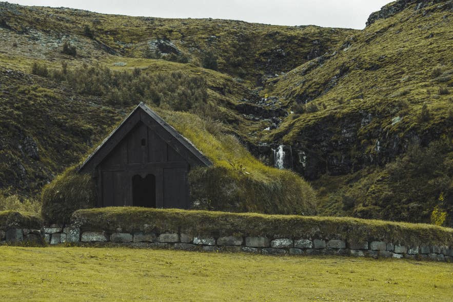 Turf-roofed longhouse at Thjodveldisbaerinn at Stong with a waterfall and green hills behind
