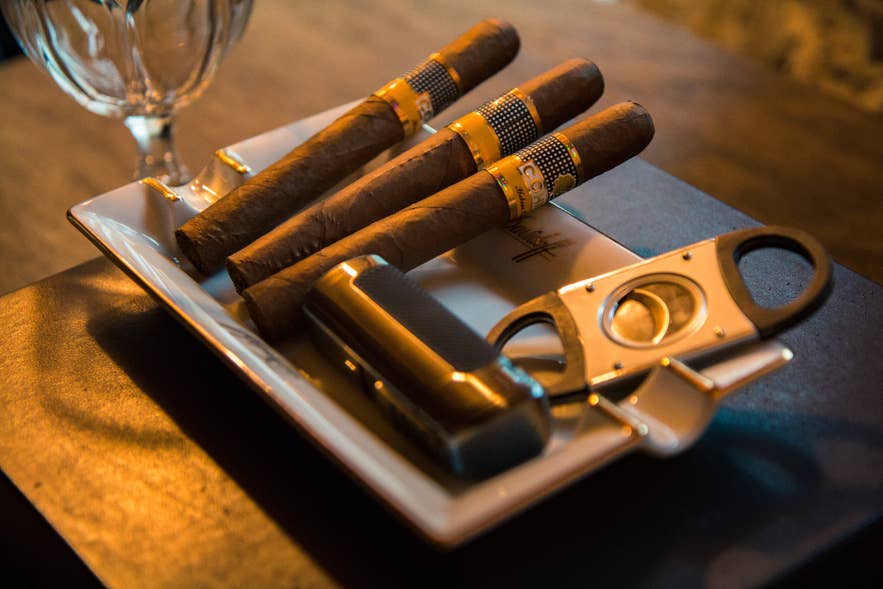 Nærbillede af luksuriøse Cohiba-cigarer på et keramisk askebæger med en cigarskærer og en lighter, placeret på en mørk træoverflade.
