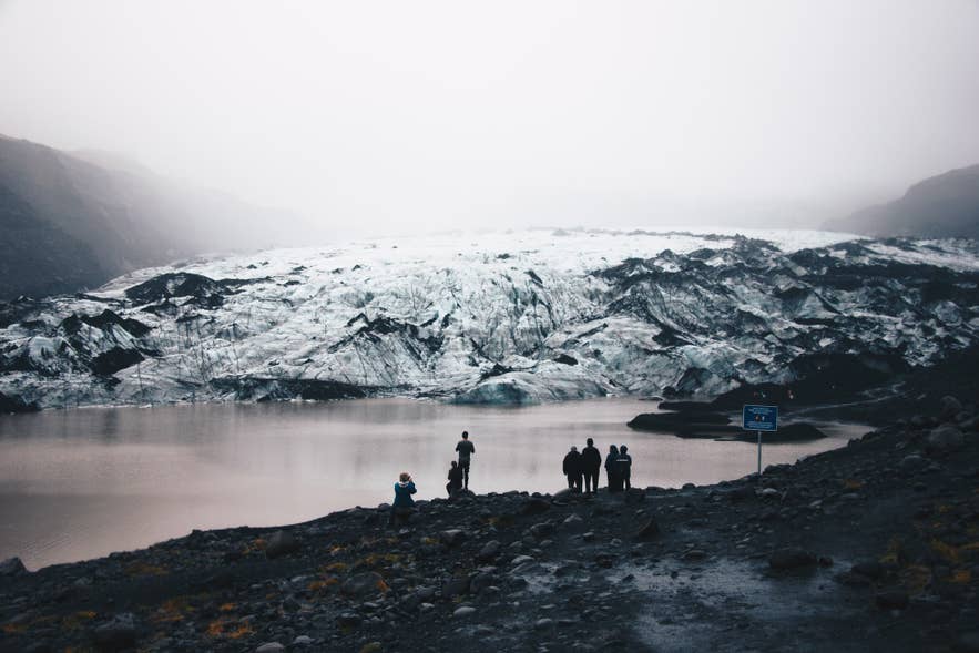 Persone davanti a un lago di fronte al ghiacciaio Myrdalsjokull, con cielo nebbioso e paesaggio ghiacciato.