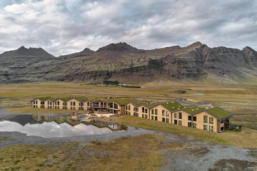 Modern Glacier Lagoon Hotel met met gras begroeide daken bij de Jokulsarlon Gletsjerlagune in IJsland, met dramatisch berglandschap en omgeven door open vlaktes en spiegelend water.