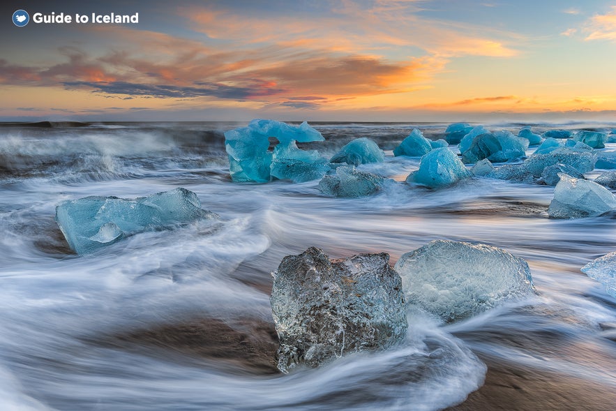 Eisstücke liegen am schwarzen Sandstrand Diamond Beach in Island bei Sonnenaufgang, mit Meereswellen, die um die kristallklaren und blauen Eisberge fließen, unter einem farbenfrohen Himmel.