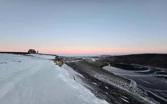 Explorador de invierno junto a un SUV en una cresta alta sobre un lago helado y un paisaje volcánico remoto.