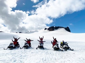 A group of people are seen sitting on snowmobiles on top of Vantajokull Glacier with their hands up in the air.