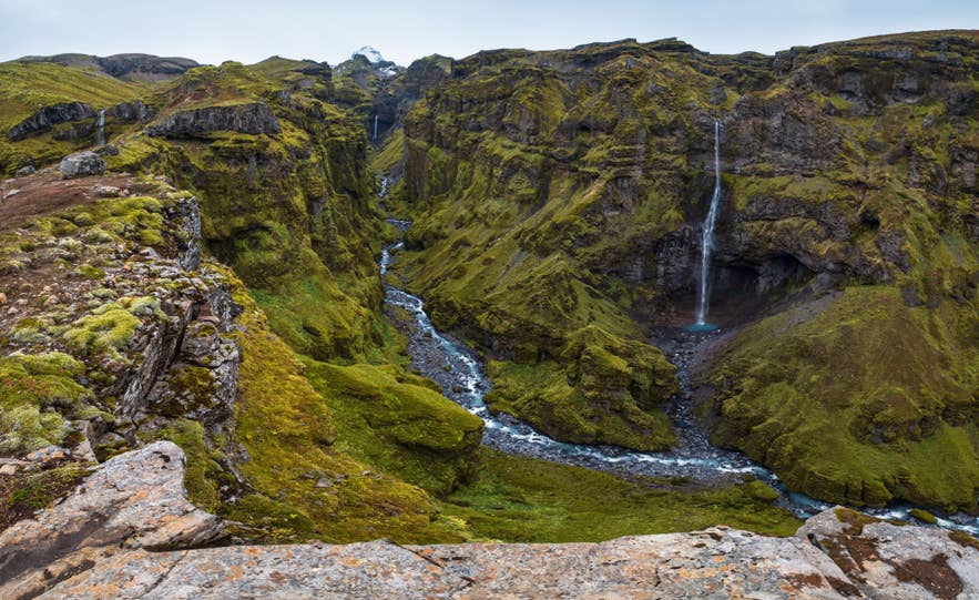 Moss-lined Mulagljufur Canyon with a winding river and a tall waterfall under a cloudy sky.
