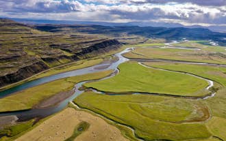 Winding river cuts through green farmland and terraced basalt cliffs in West Iceland under a dramatic sky.