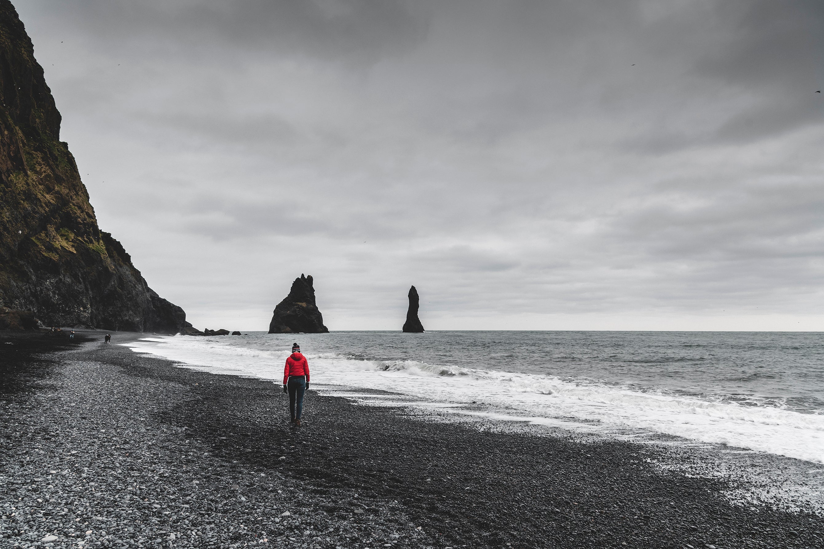 Person in red jacket walks along Reynisfjara Black Sand Beach in South Iceland with sea stacks and cliffs under a cloudy sky.