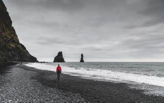 Person in red jacket walks along Reynisfjara Black Sand Beach in South Iceland with sea stacks and cliffs under a cloudy sky.