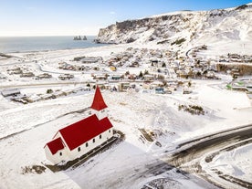 Snow-covered town of Vik in South Iceland with red-roof church in front, sea stacks in the distance, and cliffs rising behind.