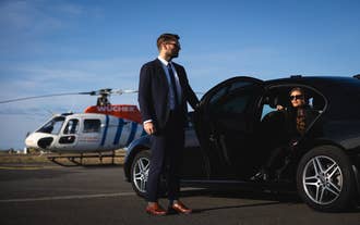 Chauffeur in a navy suit opens a car door for a woman as a helicopter waits nearby on an airport tarmac.