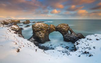 Gatklettur, den naturlige steinbuen, reiser seg majestetisk langs Islands kyst, omgitt av snø, mens bølgene slår mot klippene under.
