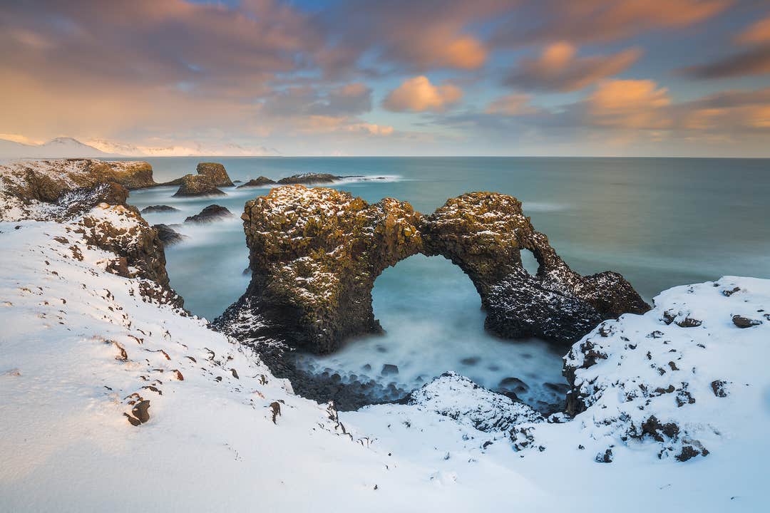 Gatklettur Natural Rock Arch stands tall on Iceland’s coastline, surrounded by snow, as waves crash below.