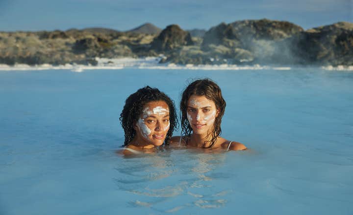 หญิงสาวสองคนพอกหน้าด้วยโคลนซิลิกา ผ่อนคลายในน้ำสีฟ้าของทะเลสาบน้ำพุร้อนที่ล้อมรอบด้วยหินลาวา