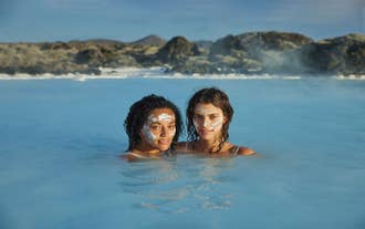 Twee vrouwen met silica-moddermaskers ontspannen zich in het rustgevende blauwe water van een geothermische lagune, omringd door lavarotsen.