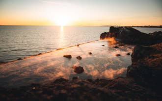 Auringonlasku Sky Lagoon -kylpylän geotermisen reunattoman infinity-altaan yllä Reykjavíkin lähellä. Kylpylän vedestä nousee höyryä, ja taustalla näkyy valtameri.