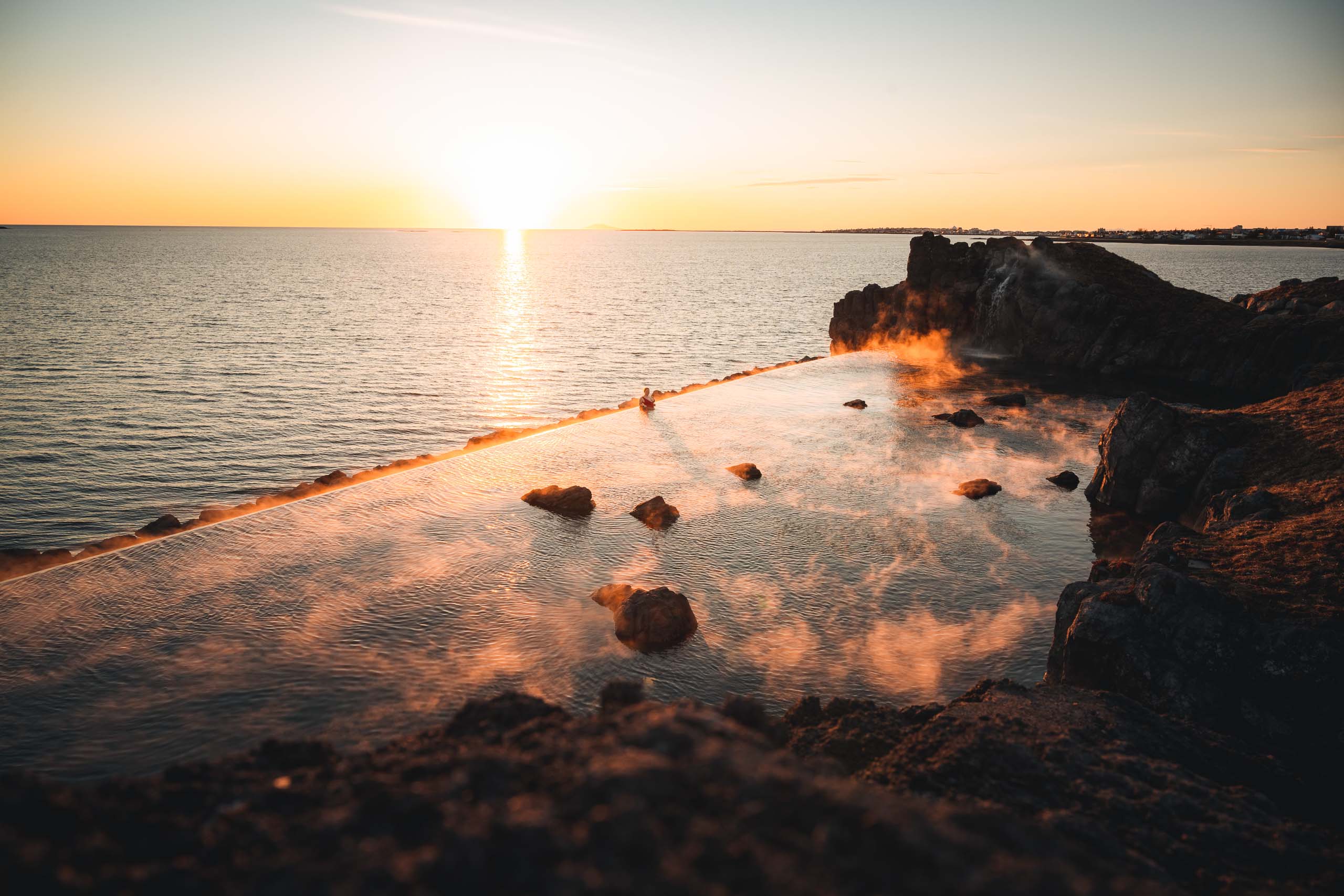 Coucher de soleil sur la piscine à débordement géothermique du Sky Lagoon près de Reykjavik, avec de la vapeur qui s’élève et une vue sur l’océan en arrière-plan.