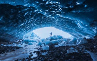 En person står inne i en blå isgrotta vid Katla på Island, omgiven av glittrande isväggar och frusna stenblock.