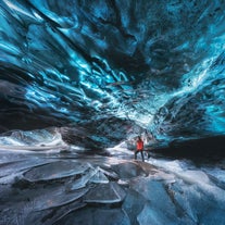 A person in red jacket walks beneath vivid blue ice cave ceiling above cracked, frozen floor in Vatnajokull National Park.