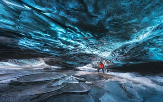 En person i rød jakke går under det blå taket i en isgrotte over det sprukne, frosne gulvet i Vatnajokull nasjonalpark.