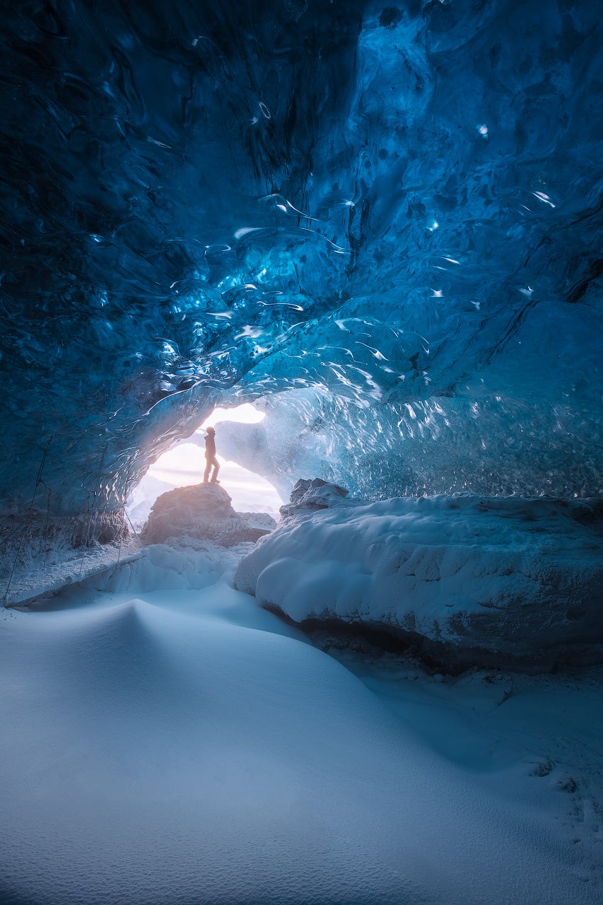 En upptäcktsresande står vid den glödande ingången till en djupblå isgrotta i Vatnajokull nationalpark, omgiven av frostväggar.