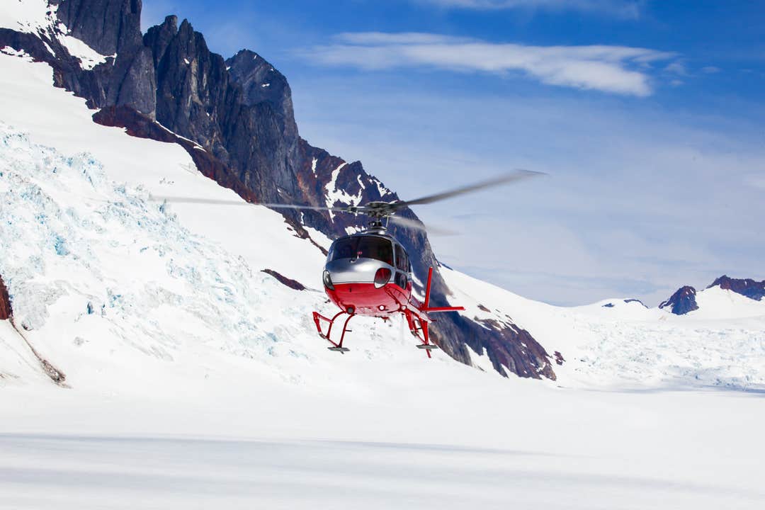 Ein rot-weißer Hubschrauber fliegt über einen verschneiten Gletscher nahe zerklüfteter Berge unter klarem, blauem Himmel.