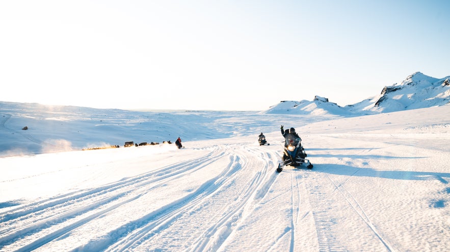 Group of snowmobilers riding across a snowy glacier in Iceland under clear blue skies, surrounded by winter landscapes and distant mountains.