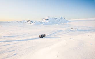 Superjeep ajaa laajalla, lumisella jäätiköllä Islannissa; taustalla vuorenhuiput ja kirkas taivas.
