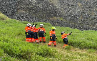 Personas en cuatrimotos con trajes naranjas y negros, tomando fotos y señalando lugares de interés en un valle verde junto a formaciones de roca basáltica.