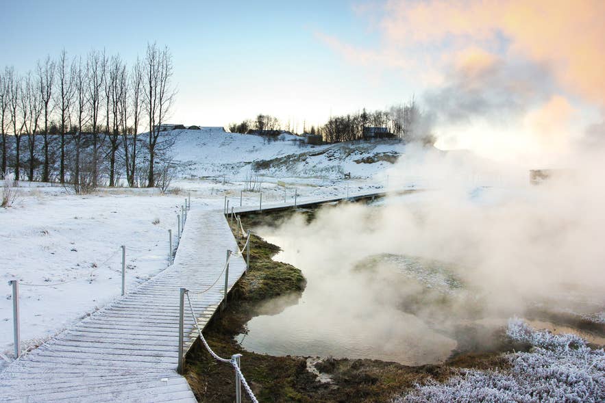 Snow-covered wooden boardwalk with rope railings beside a steaming hot spring at the Secret Lagoon