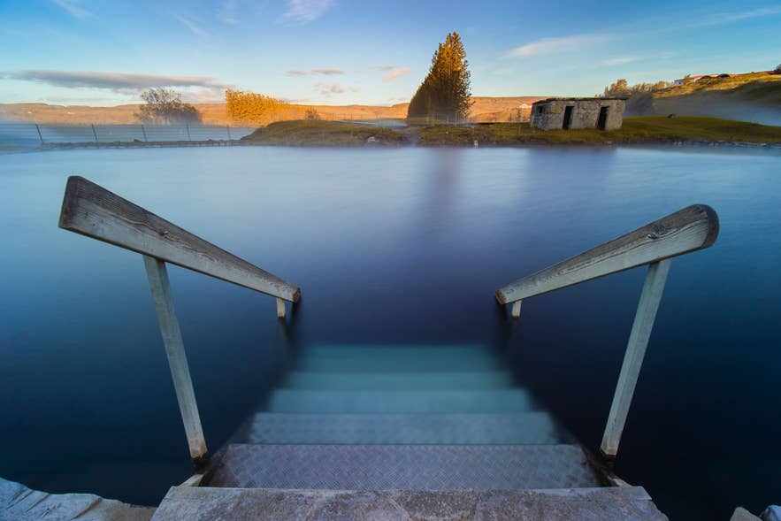 Metal steps with wooden handrails leading into the Secret Lagoon's main pool, with the old stone building beyond