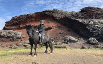 Un cavalier monté sur un cheval islandais traverse un spectaculaire paysage volcanique, entouré de roches de lave rouge éclatantes sous un ciel bleu lumineux.