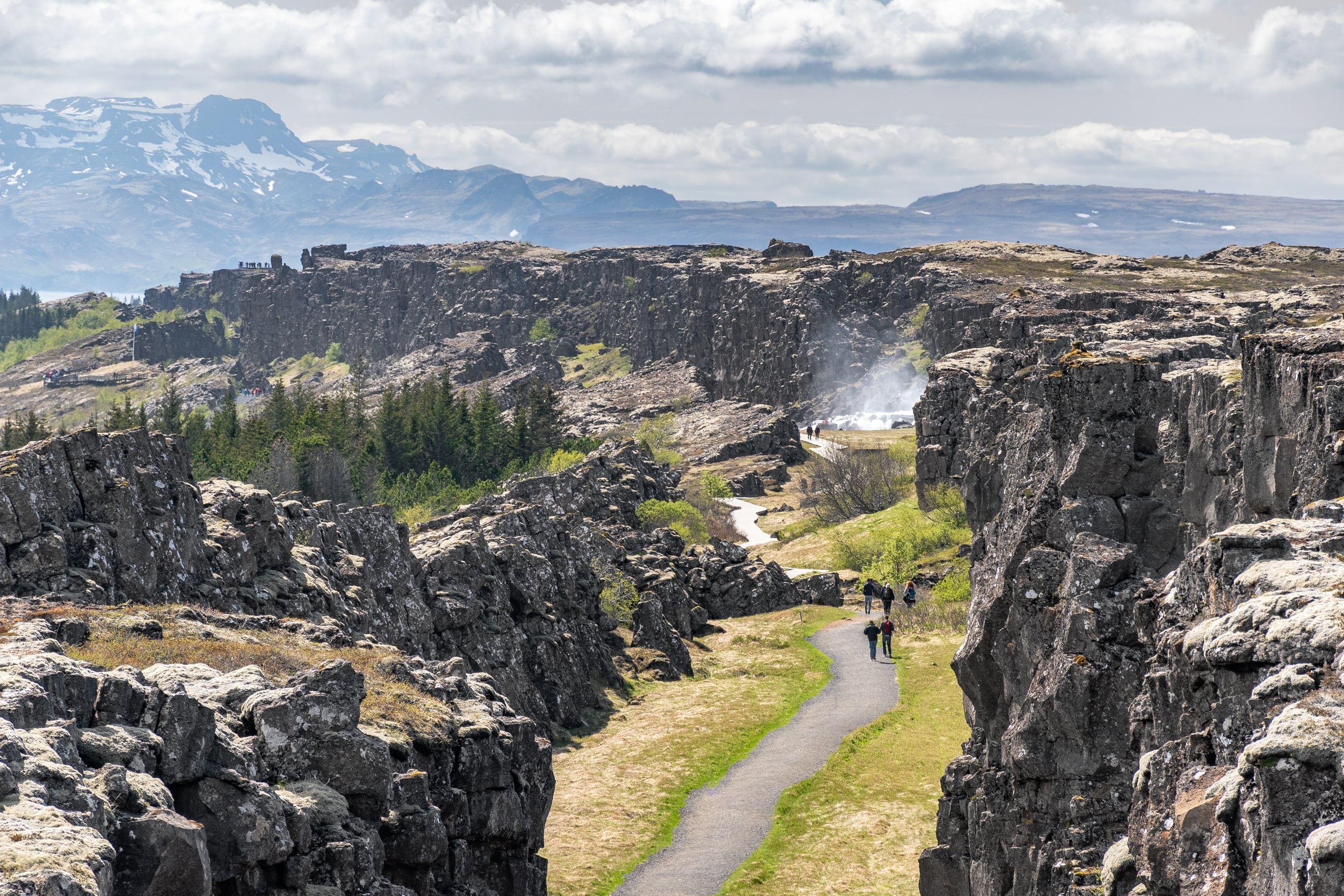 Menschen, die durch die Almannagja-Schlucht im Thingvellir-Nationalpark spazieren, mit Lavafelsen, Grünflächen und Dampf, der in der Nähe eines Geysirs aufsteigt.