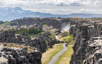 People walking through the Almannagja Gorge at Thingvellir National Park, with lava cliffs, greenery, and steam rising near a geyser.