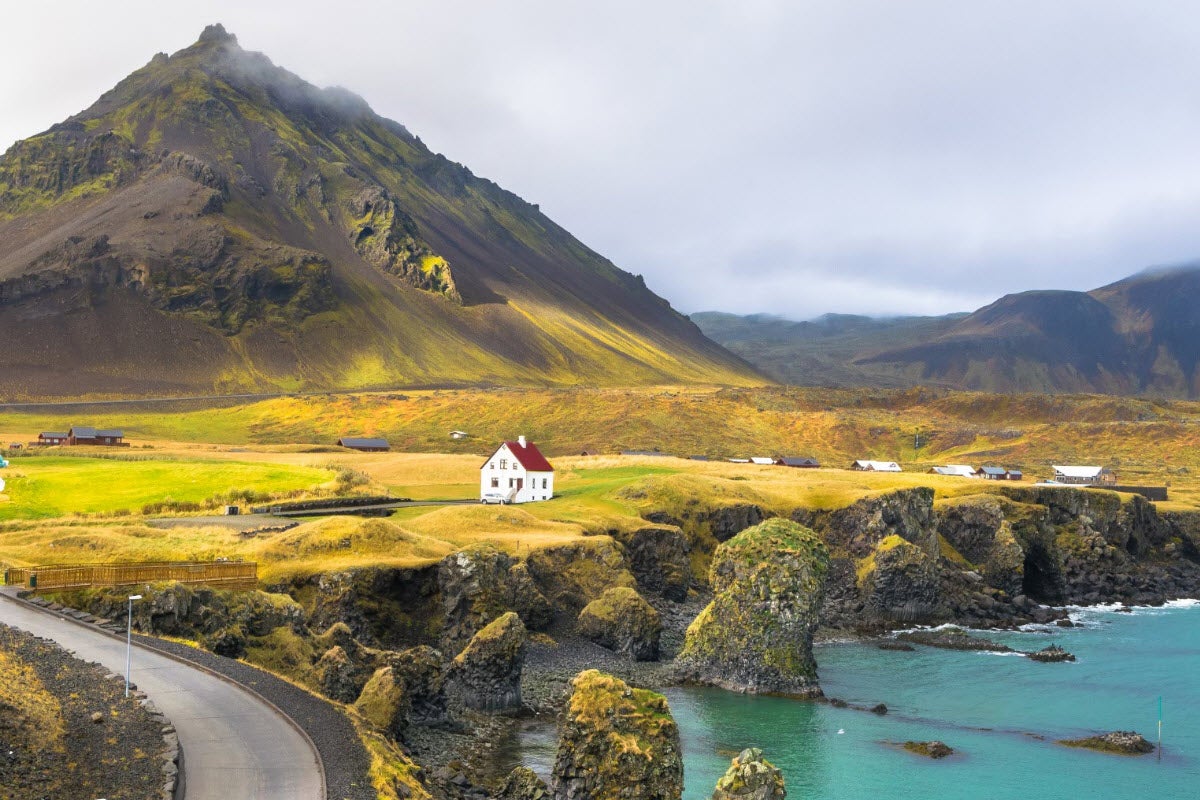 White house on Arnarstapi coastline with sea cliffs and mossy mountain in Snaefellsnes Peninsula.