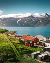 Scenic view of Djupavik village with colorful buildings by a turquoise fjord, backed by snow-streaked mountains and low clouds.