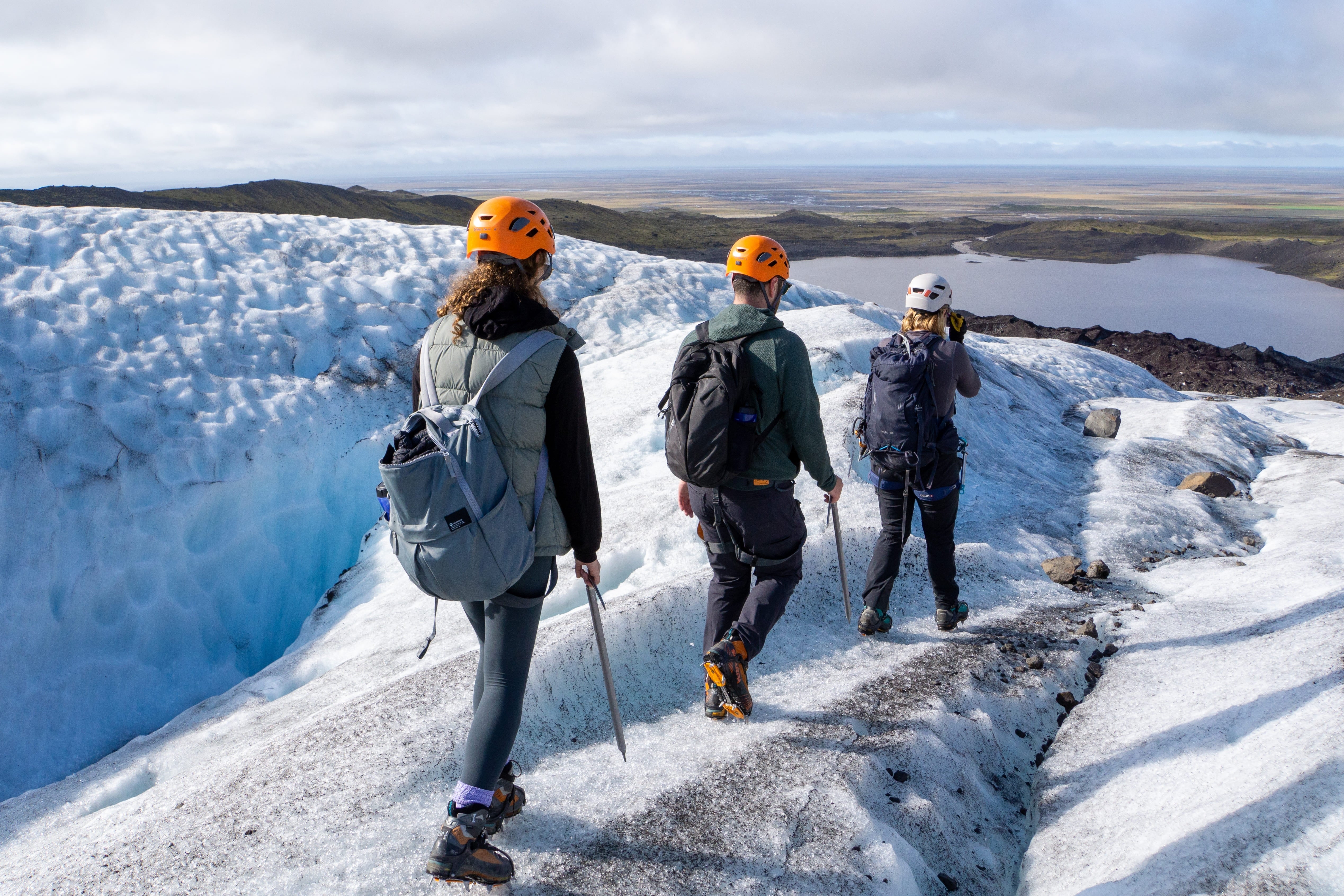 Three explorers hike across a vast Icelandic glacier, equipped with helmets and crampons.