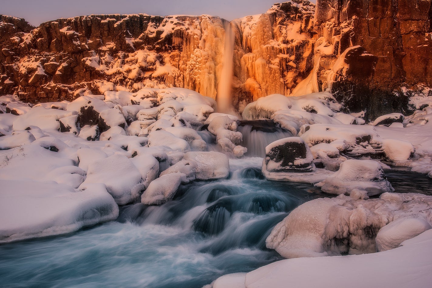 Frozen Oxararfoss Waterfall in Thingvellir National Park glows at sunset with icy rocks and rushing water.