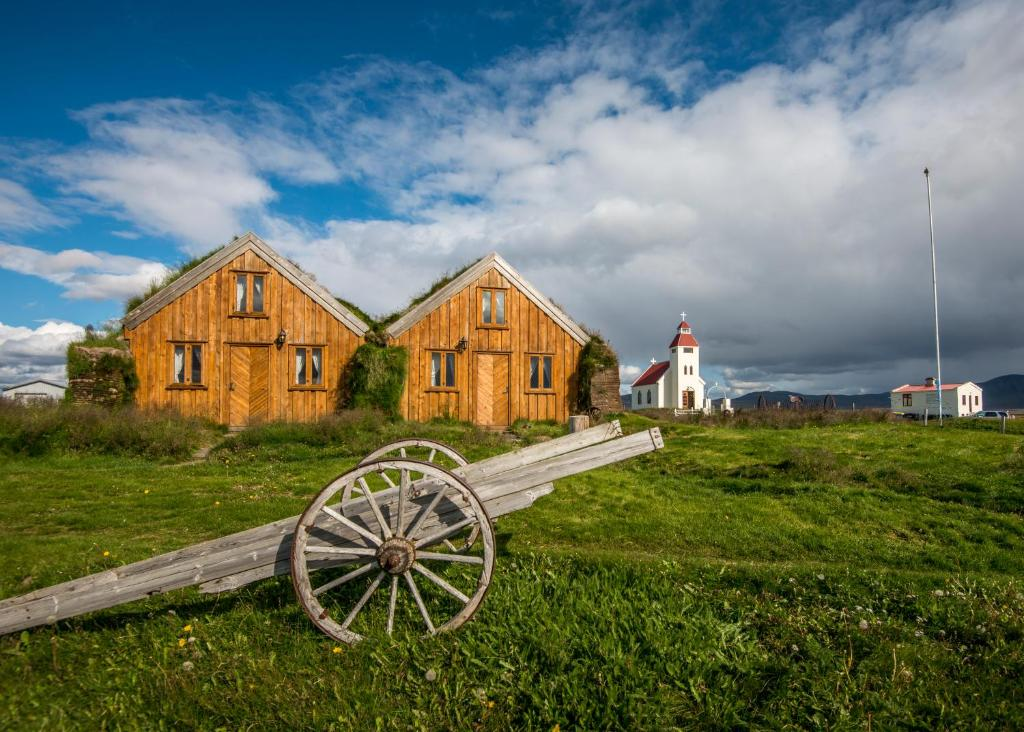 Three wooden turf-roofed cabins with a rustic cart in front and a white church in the distance under a bright blue sky in Modrud