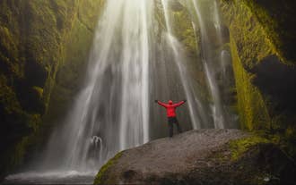 A hiker in a red jacket stands by the hidden Gljufrabui Waterfall, surrounded by mossy cliffs in South Iceland.