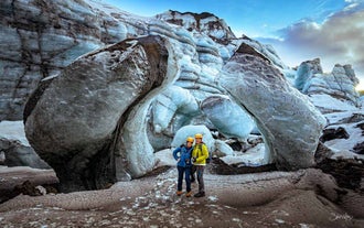 Two visitors pose at the base of a glacier surrounded by blue and black ice formations.
