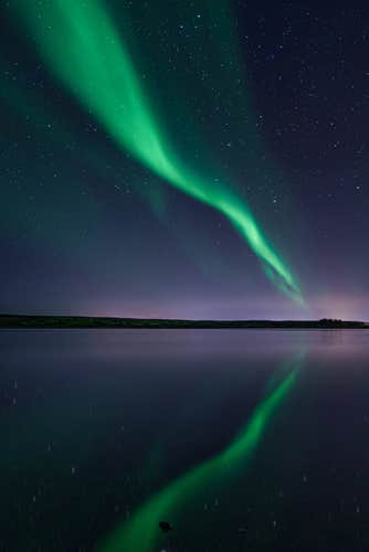 A green streak from the aurora borealis lights up the winter night sky and reflects onto the water below.