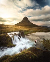 Woman wearing bobble hat standing looking at Kirkjufell mountain and waterfall.