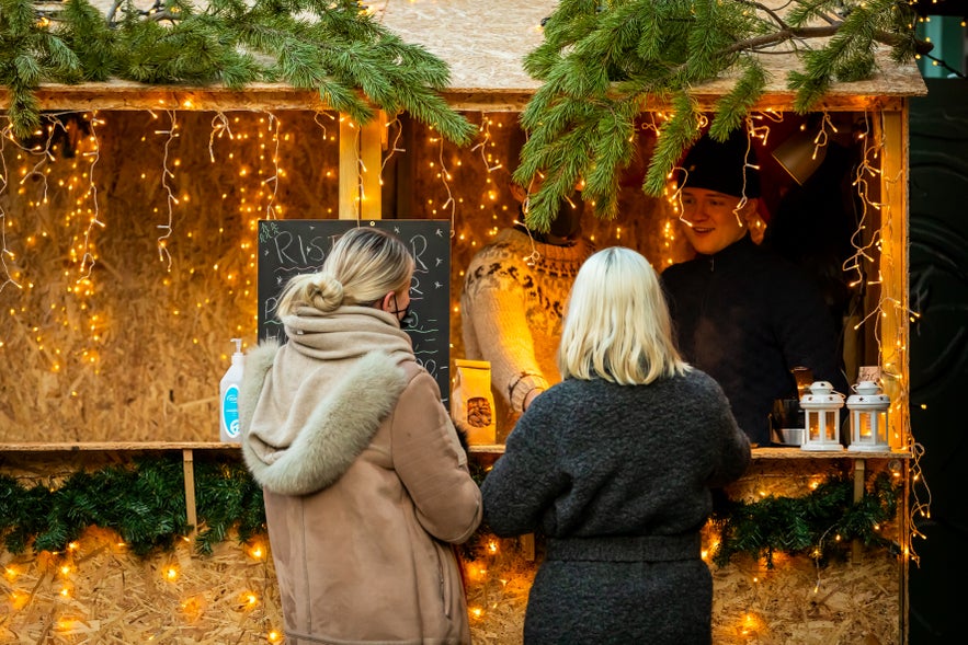 Two women on a Reykjavík Christmas walking tour visit a festive wooden stall at the city’s holiday market, warmly lit with fairy lights and evergreen decorations.