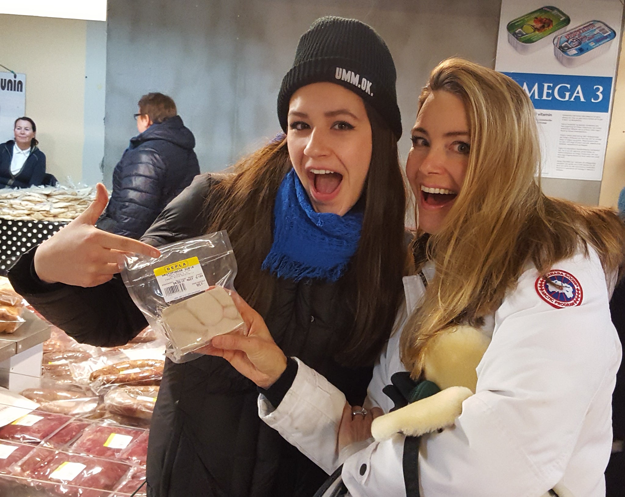 Two women wearing hats. One is pointing at some traditional Icelandic food.